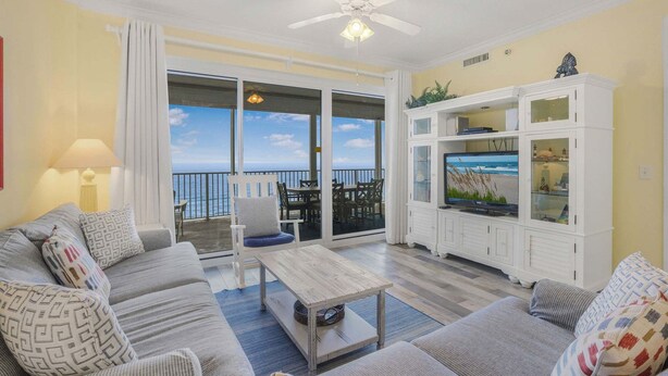 A living room with a sofa, coffee table, and TV stand, a balcony with a view, and a ceiling fan. - Regency Isle 711  2 BR 2 5 BA  sleep 8  gulf-front (Orange Beach)