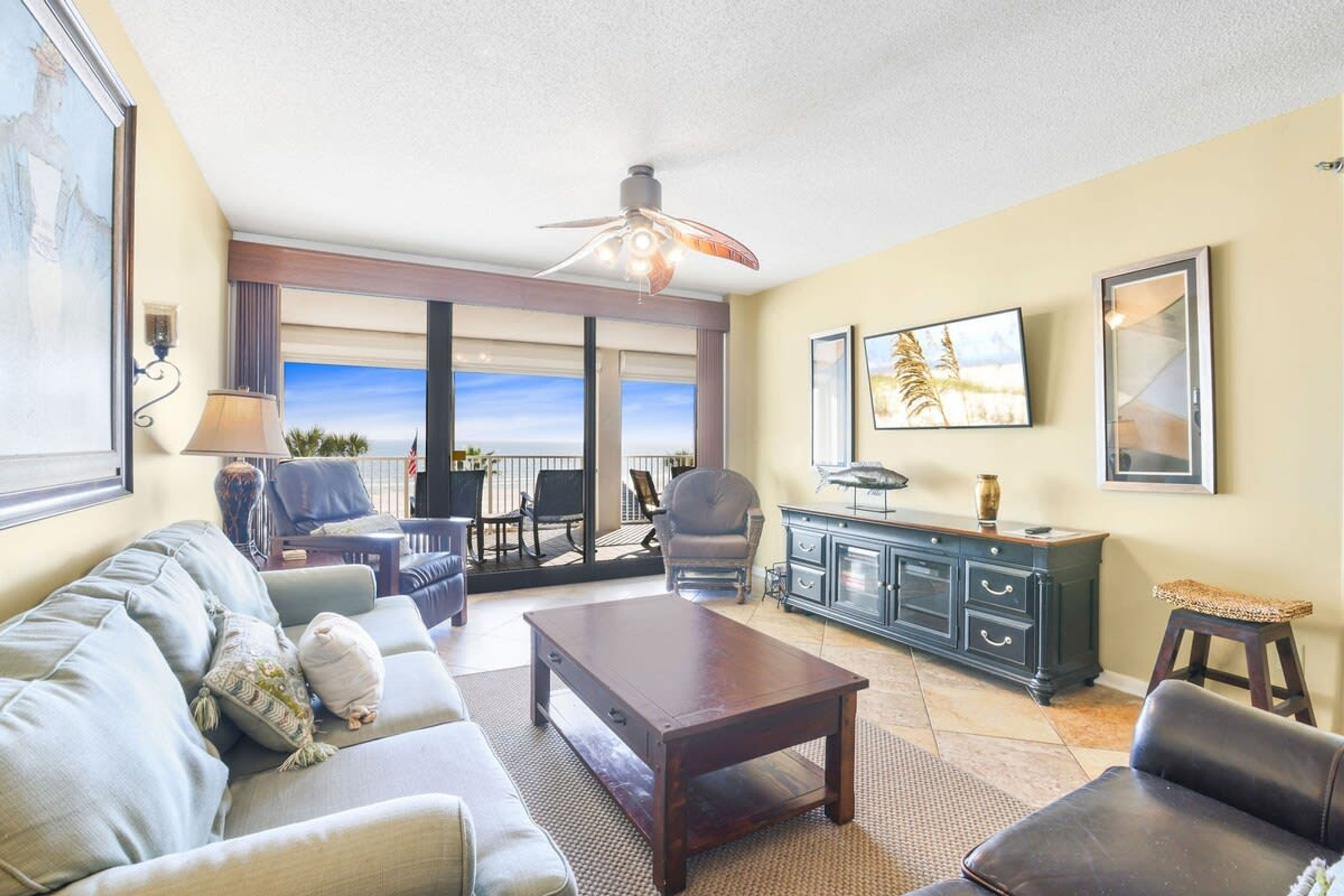 A spacious living room with a large window offering a view of the ocean, a ceiling fan, and a dark wooden coffee table.