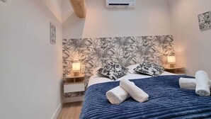 Une chambre avec un puits de lumière, un lit recouvert d’une couverture bleue, deux tables de chevet avec des lampes et un mur orné d’un motif de feuilles.