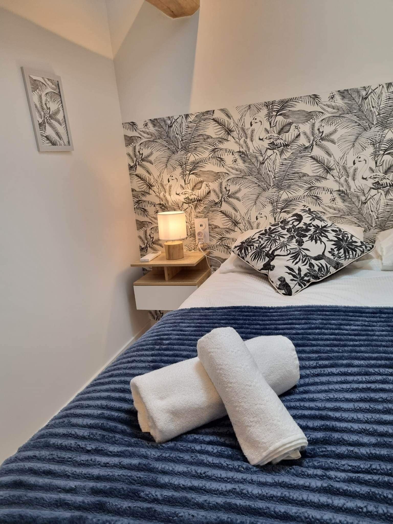 En pænt redt seng med et blåt stribet lagen, en hvid pude med et sort blomstermønster og en sengetavle i træ med en lampe.