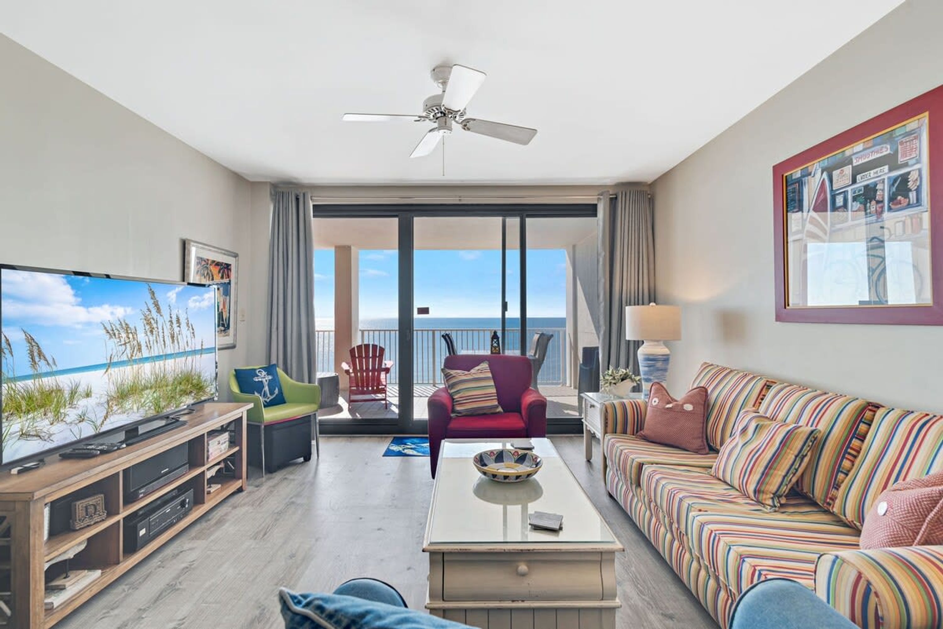 A spacious living room with a large flat-screen TV, a ceiling fan, a sofa, a coffee table, and a balcony with a view of the ocean.