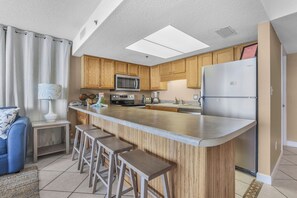 A modern kitchen with wooden cabinets, a stainless steel refrigerator, a microwave, and a built-in island with bar stools. - Phoenix II 2101  3 bedrooms  Amenities Galore  Beachfront (Orange Beach)