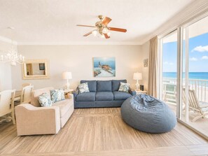Ein Zimmer mit Deckenventilator, einem großen Fenster mit Blick auf den Strand, einem blauen Sofa, einem beigen Sessel und einem Sitzsack.