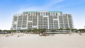A multi-story hotel with a flat roof, located on a sandy beach with palm trees in the foreground. - Regency Isle  Beachfront Bliss   Amazing Views  2 bedrooms (Orange Beach)