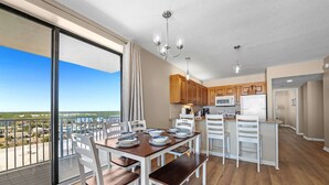 A modern kitchen with wooden cabinets, a dining area with white chairs and a wooden table, and a balcony with a city view. - Shoalwater 906  3 3  Gulf-front  CORNER unit (Orange Beach)
