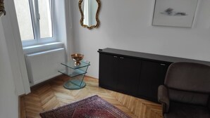 Une pièce dotée d’un parquet en bois disposé en motif «point de Hongrie», d’un fauteuil en cuir marron foncé, d’une table en verre surmontée d’un saladier, d’un meuble noir et d’un grand tapis à motifs.