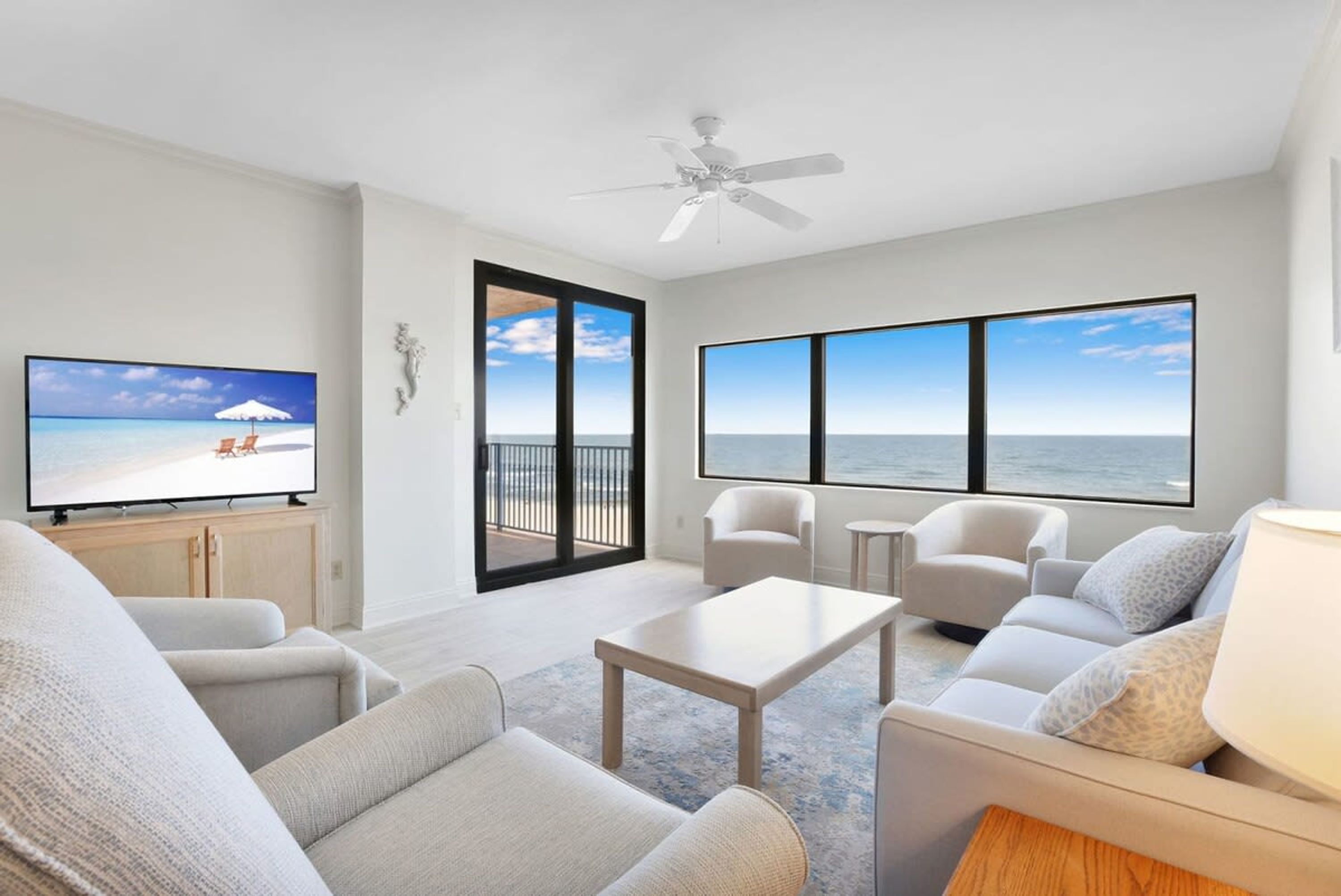 A spacious living room with a large TV, comfortable seating, a ceiling fan, and a balcony with a view of the ocean.