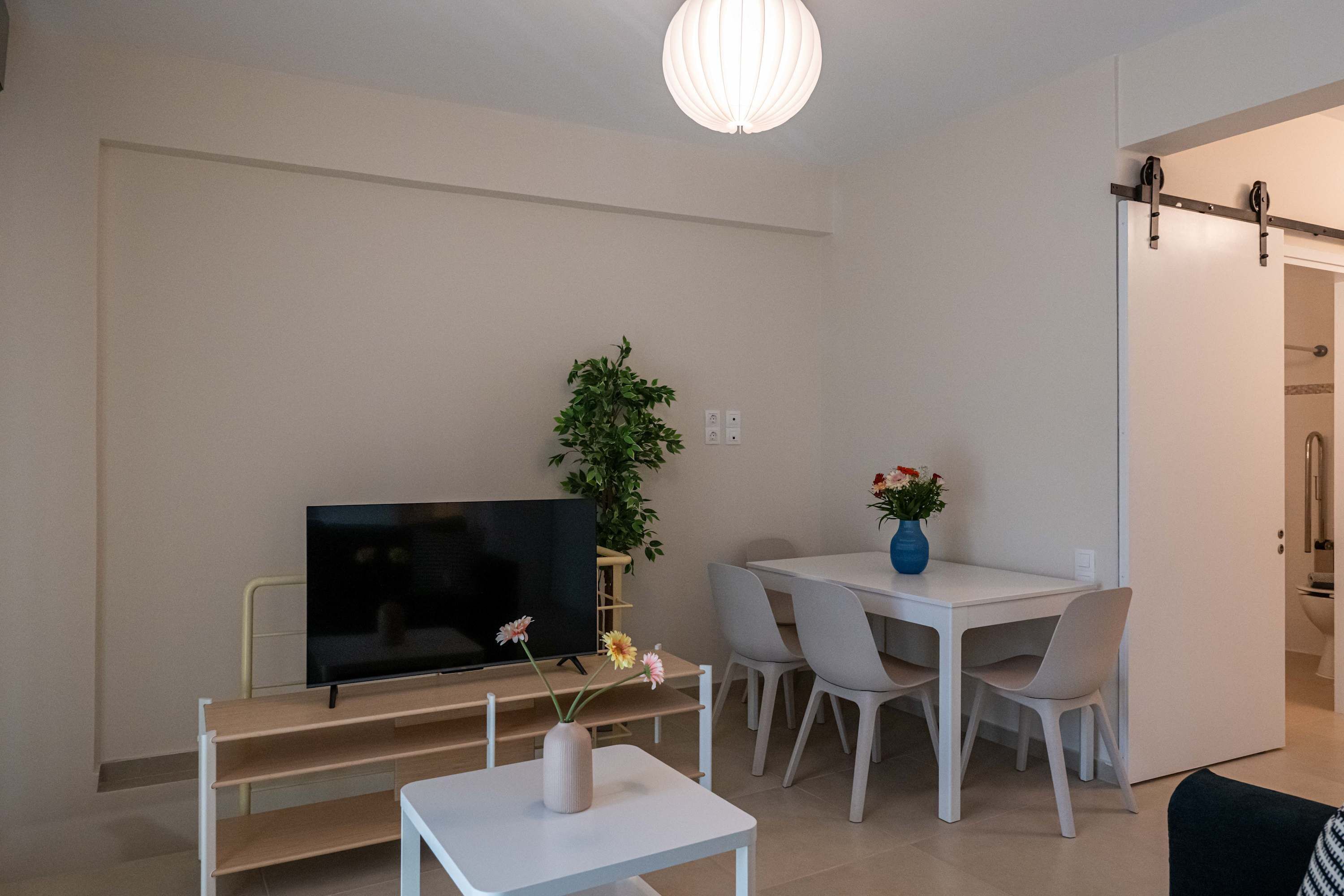 A modern living room with a flat-screen TV, a white coffee table, a dining table with chairs, a potted plant, and a sliding door leading to another room.