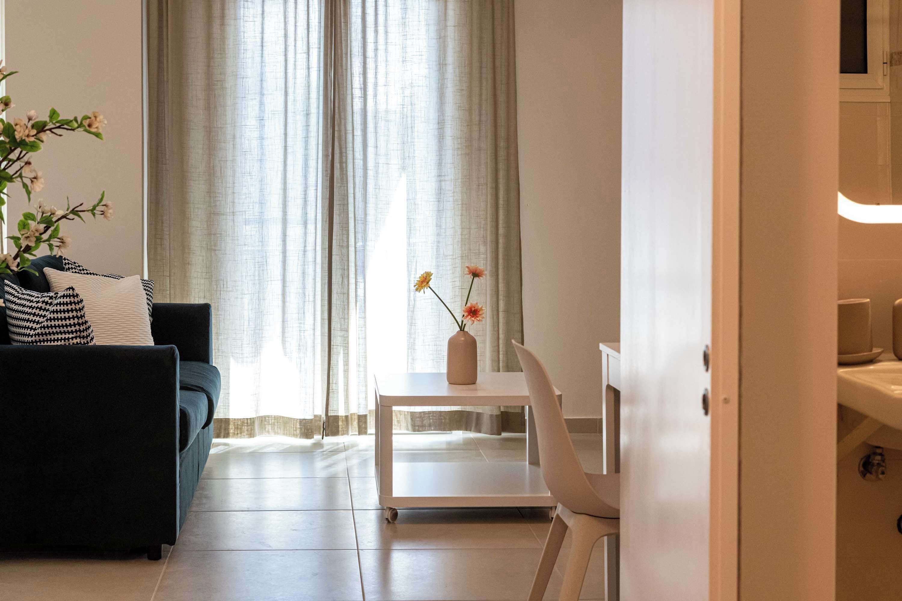 A modern living room with a sofa, a small white table with a vase of flowers, and a view of a kitchen area.