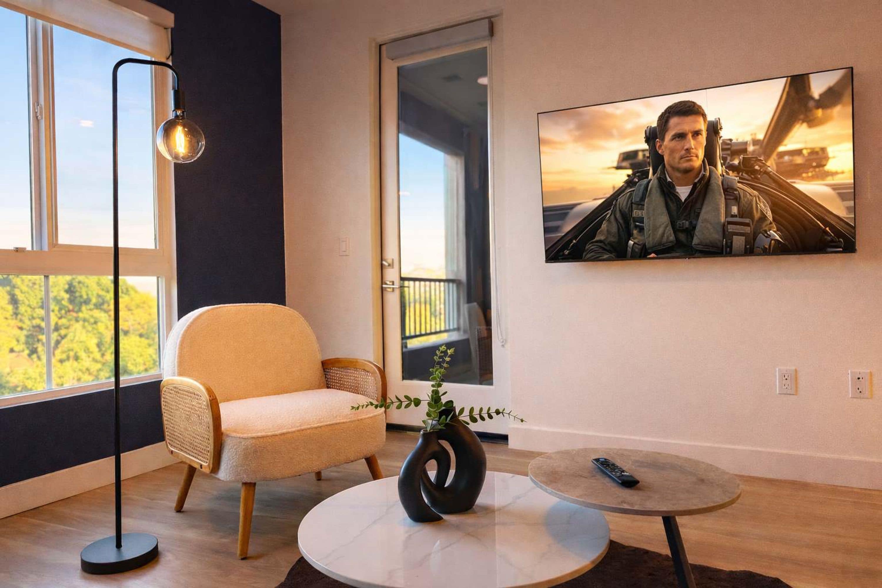 A modern living room with a large window, a comfortable chair, a round coffee table, and a flat-screen TV mounted on the wall.
