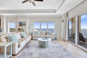 A spacious living room with a large window offering a view of the ocean, a ceiling fan, and a round coffee table. - Beachfront Escape  Sleeps 10  Incredible Views (Orange Beach)