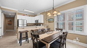 A kitchen with white cabinets, a stainless steel refrigerator, and a microwave. There is a dining area with a wooden table and chairs, and a window with blinds. - Island Royale- Beachfront Penthouse (Gulf Shores)