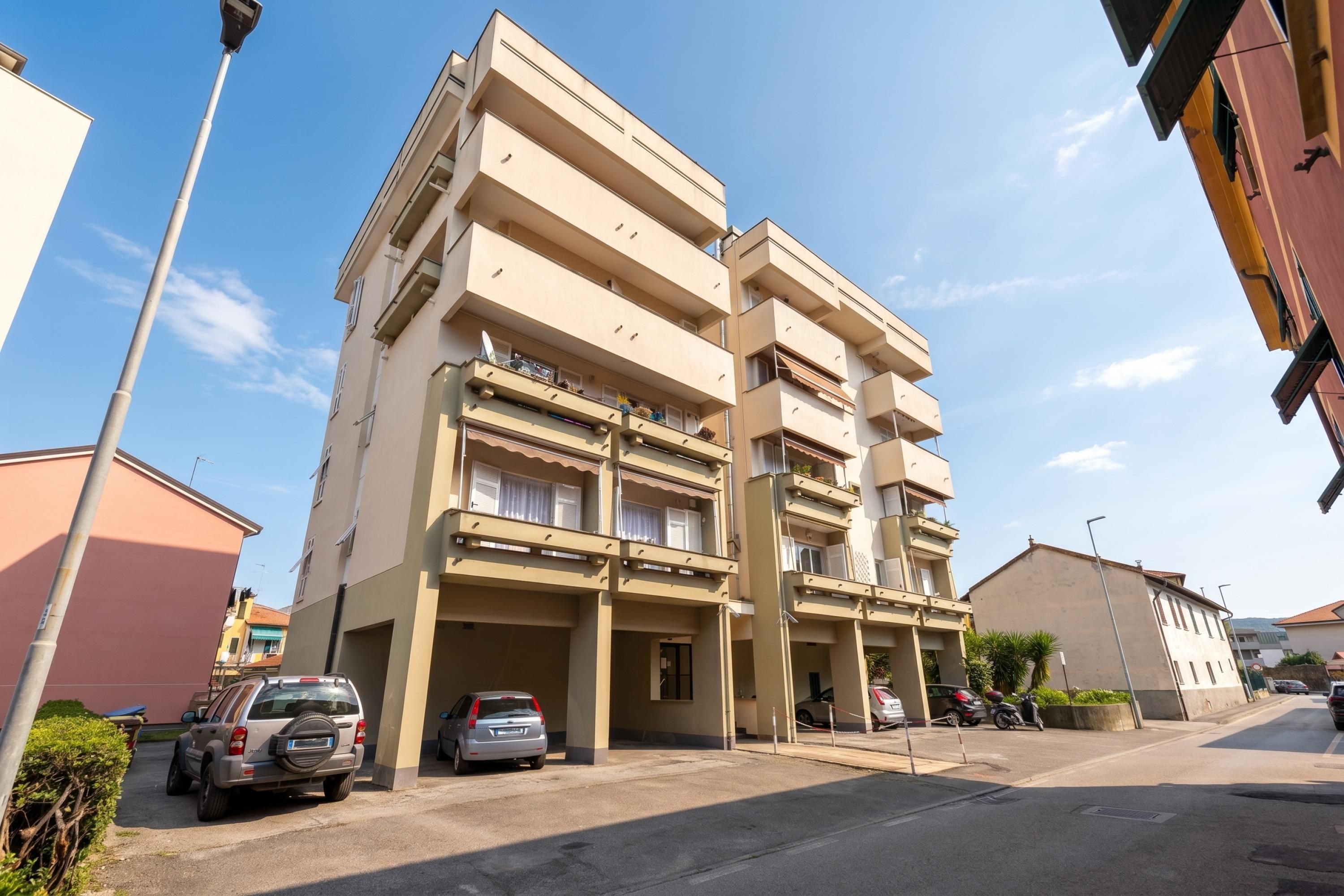 Un edificio residencial de varios pisos con balcones, coches estacionados y un cielo despejado.