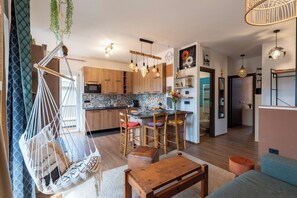 Una cocina moderna con armarios de madera, un rincón comedor con taburetes de bar, una silla colgante de macramé e iluminación colgante.