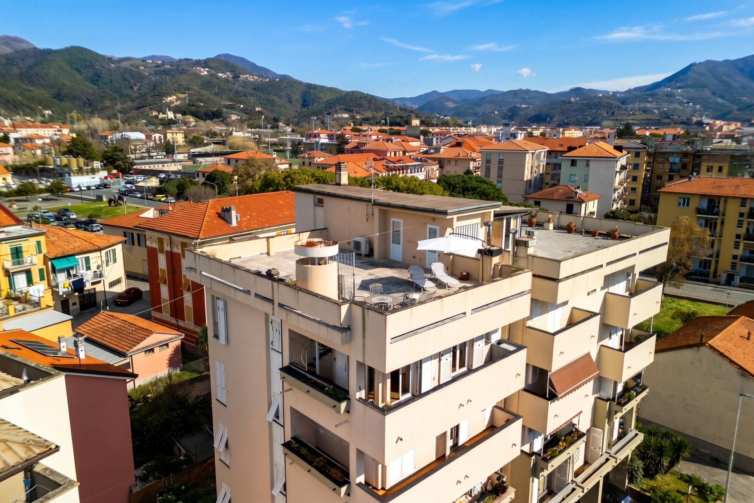 Un immeuble à plusieurs étages doté de balcons, d’une terrasse sur le toit et offrant une vue sur une ville et des montagnes en arrière-plan.