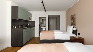 A compact hotel room with a kitchenette, a bed with a beige blanket, a bedside table with a lamp, and a painting on the wall. - Limehome Düsseldorf Kö (Düsseldorf)