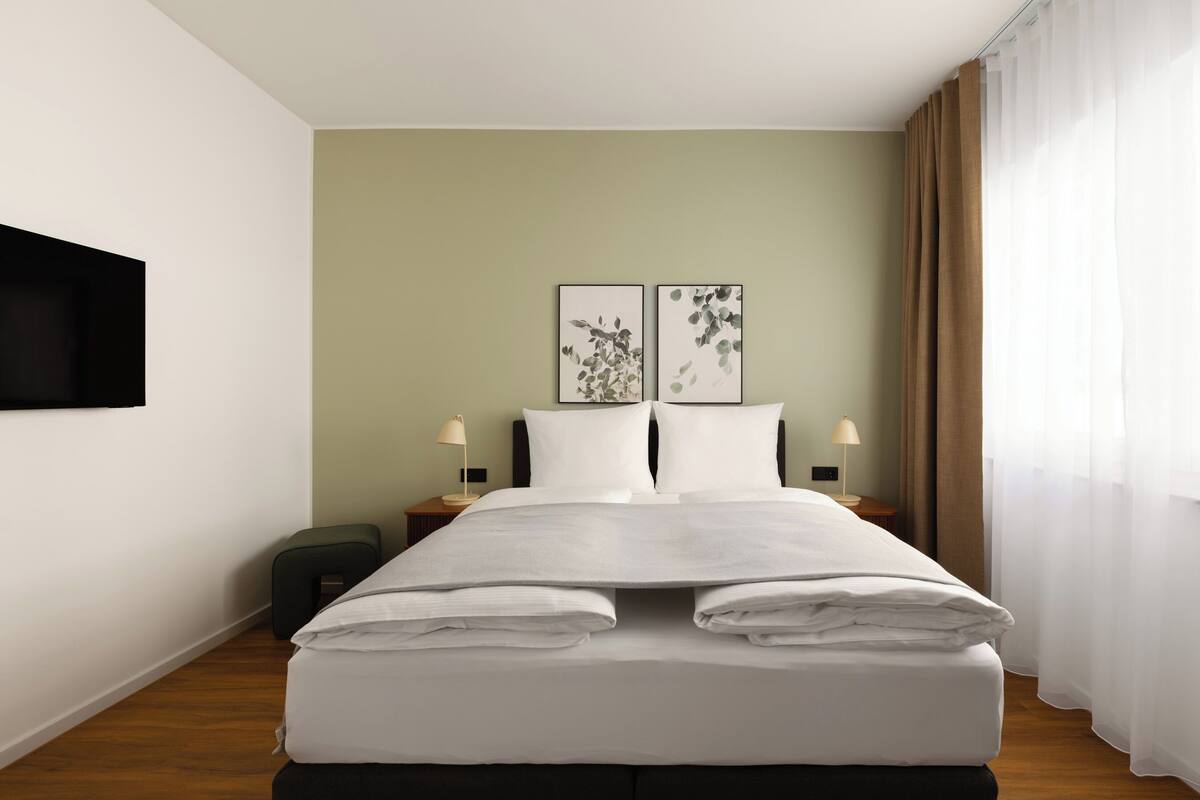 Ein Hotelzimmer mit Bett, zwei Nachttischen, einem an der Wand montierten Fernseher und Kunstwerken über dem Bett.