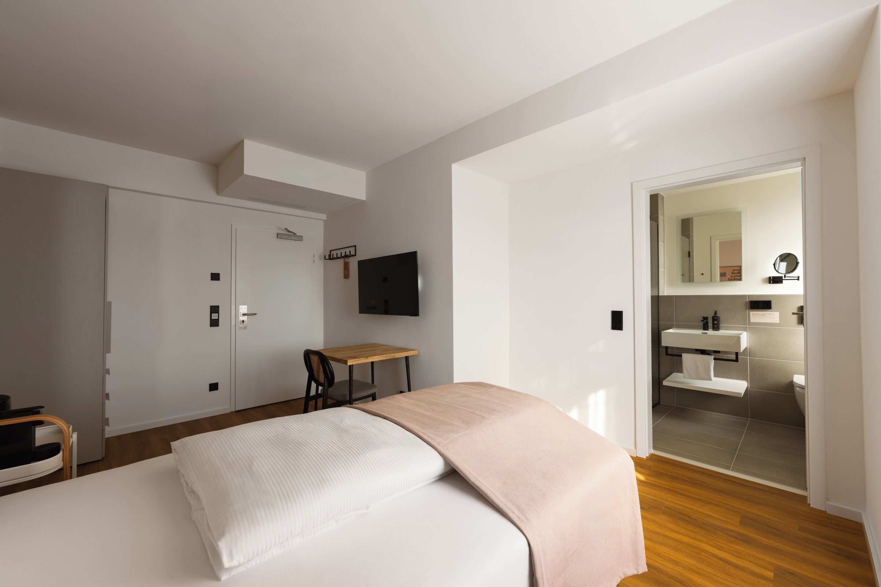 Un dormitorio con cama, escritorio, televisor, armario y baño visible a través de una puerta abierta.