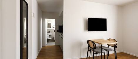 A modern interior with a wooden floor, a dining area with two chairs, a flat-screen TV mounted on the wall, and a hallway leading to a bathroom.