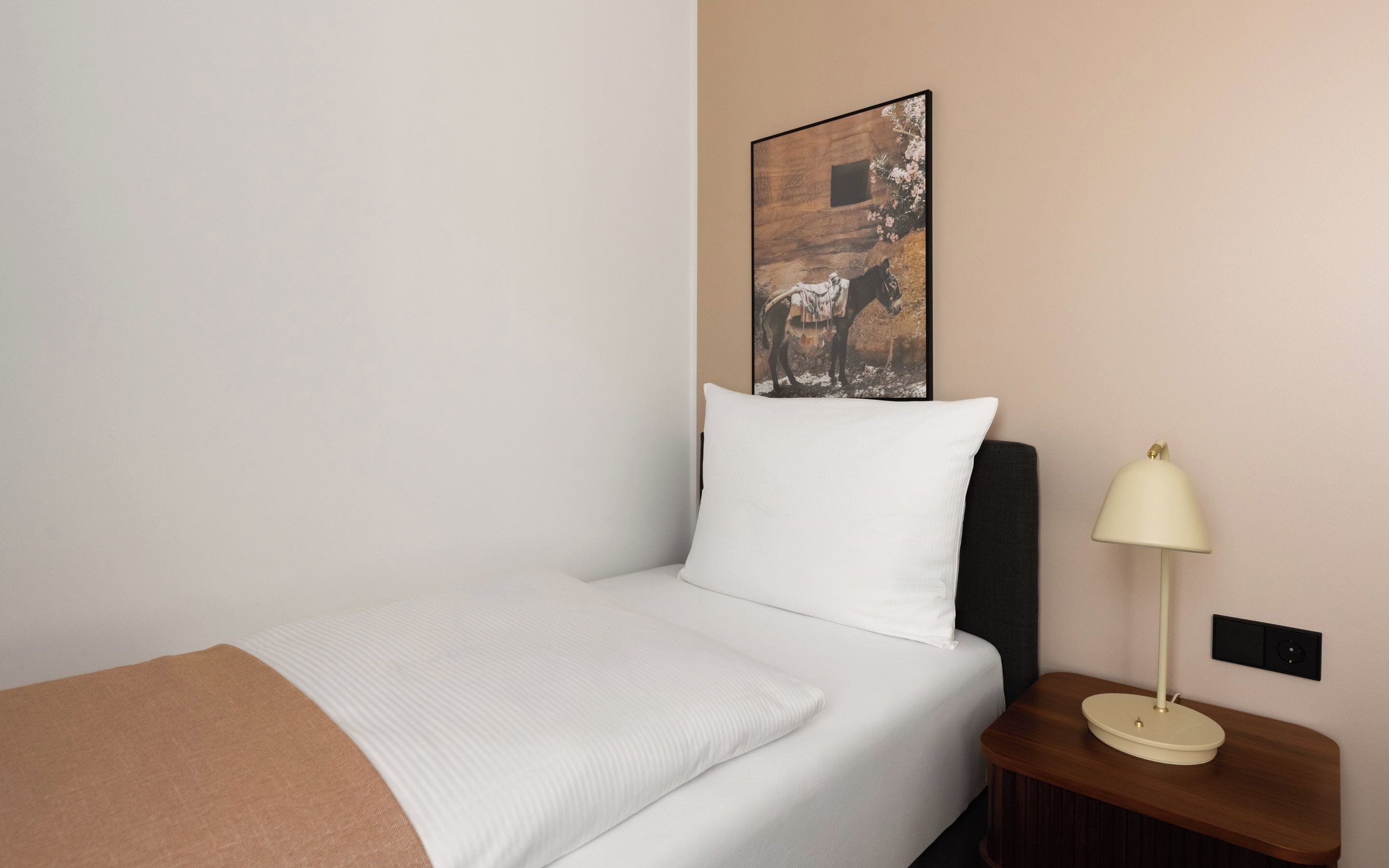 Een keurig opgemaakt bed met een wit kussen en een bruine deken, een nachtkastje met een lamp, en een aan de muur hangend schilderij van een paard.
