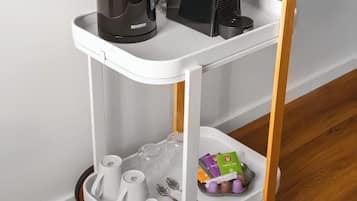 A white and wooden coffee cart with a black coffee maker, a black electric kettle, and a tray with cups and tea packets.
