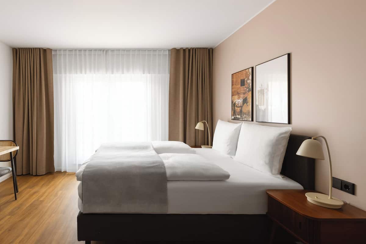 Ein Hotelzimmer mit einem großen Bett, einem Nachttisch mit Lampe, einem Sessel und einem Fenster mit Vorhängen.