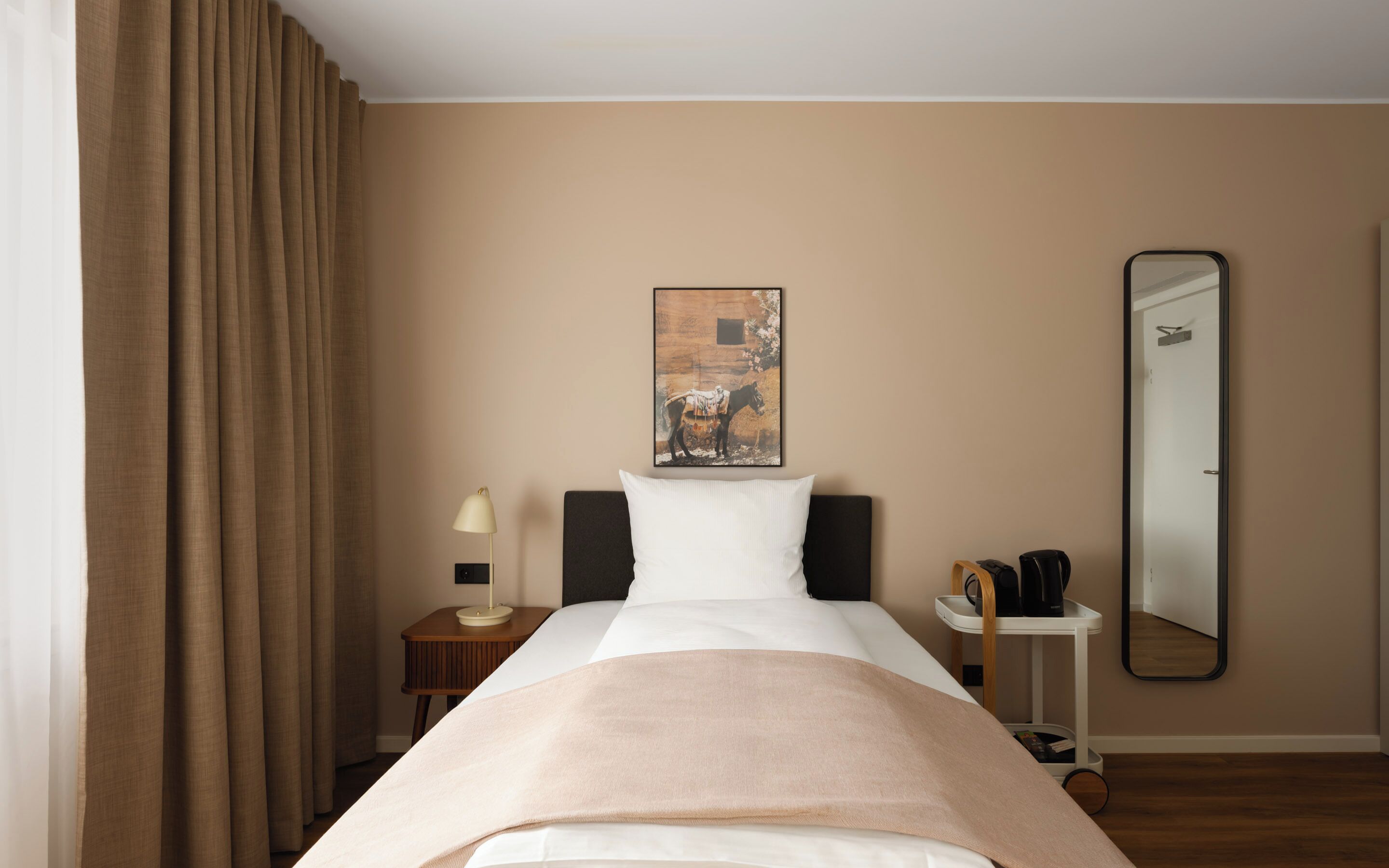 A hotel room with a bed, a nightstand, a lamp, a coffee maker, a painting, and a mirror.