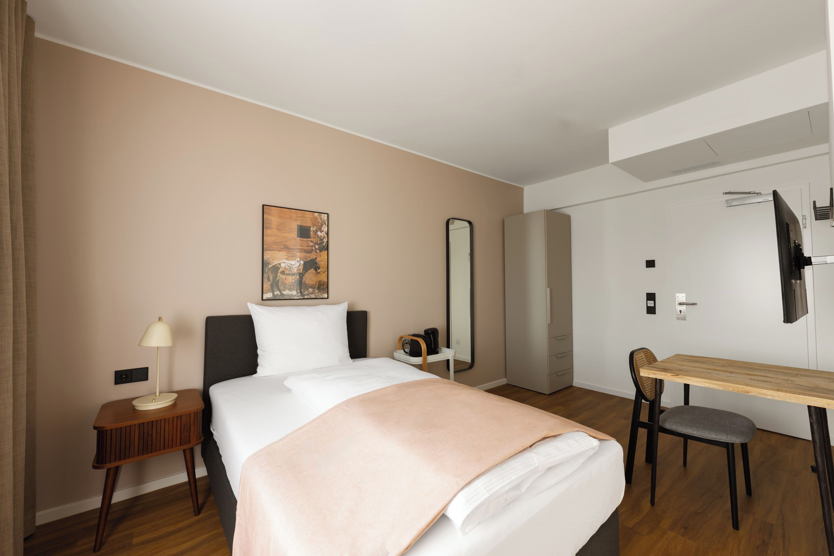 A hotel room with a bed, bedside table, lamp, chair, desk, mirror, artwork, and a wardrobe.