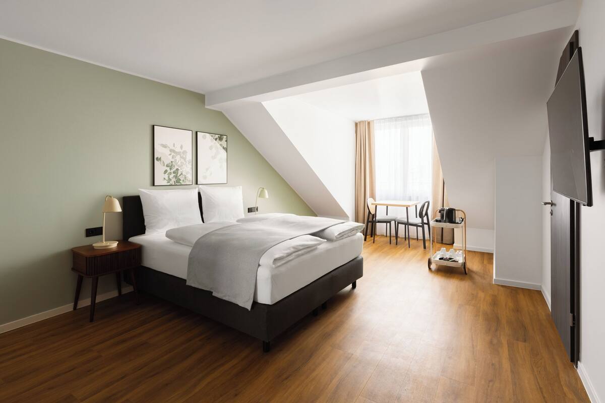 Ein modernes Schlafzimmer mit Bett, Nachttisch, Lampe und Blick auf einen Essbereich.