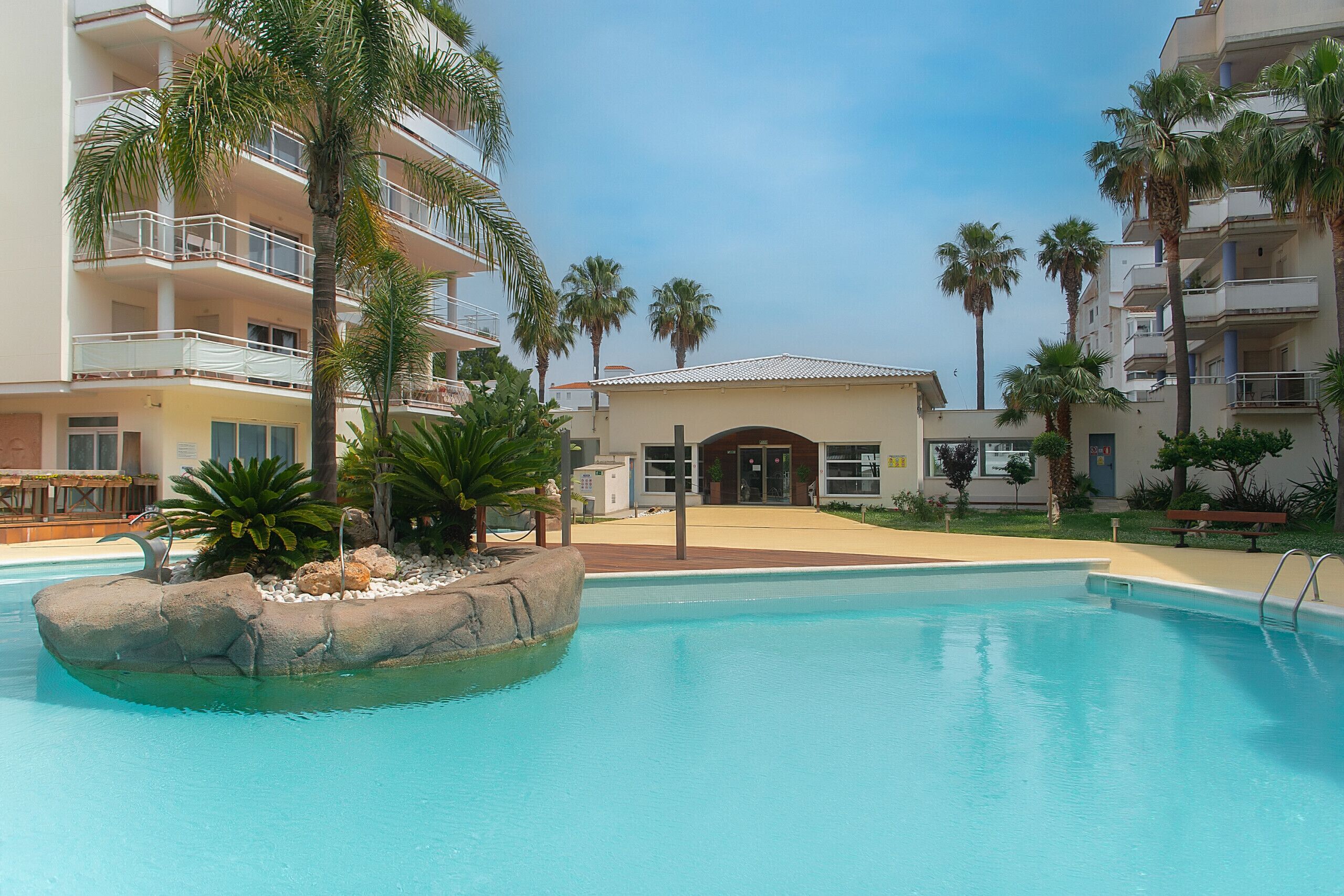 A swimming pool with a rock feature, surrounded by palm trees and a building with balconies.