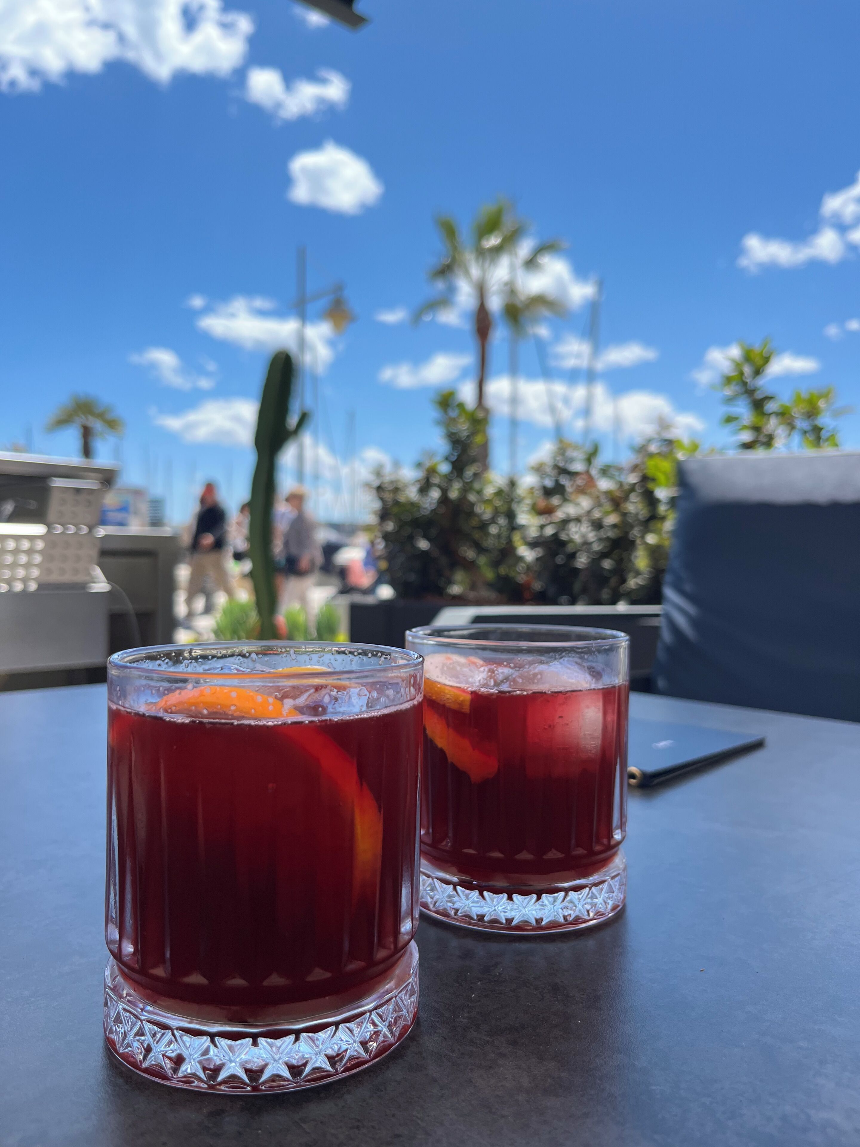 Dos vasos de bebida roja sobre una mesa, con vista despejada al cielo y palmeras al fondo.