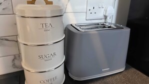 Tres recipientes apilados etiquetados 'TEA', 'SUGAR' y 'COFFEE' sobre una encimera de cocina junto a una tostadora.