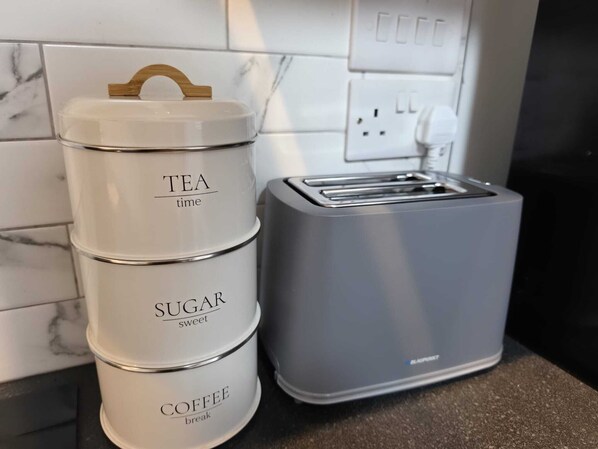 Tre staplade behållare märkta med "TE", "SOCKER" och "KAFFE" står på en köksbänk bredvid en brödrost.