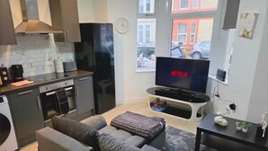 Un espacio habitable compacto con kitchenette, un sofá gris, un televisor de pantalla plana mostrando 'NETFLIX' y una ventana con vista a coches estacionados.