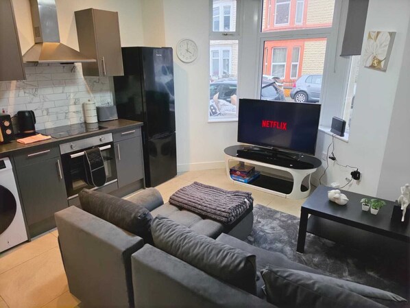 Un espacio habitable compacto con kitchenette, un sofá gris, un televisor de pantalla plana mostrando 'NETFLIX' y una ventana con vista a coches estacionados.