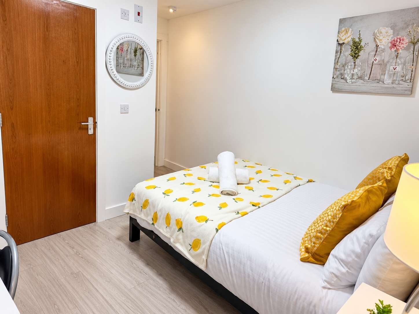A bedroom with a bed, a bedside table, a lamp, a door, and a wall decoration.