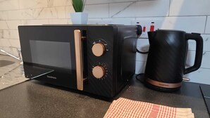 Une cuisine avec un micro-ondes noir, une bouilloire et un torchon à carreaux sur le plan de travail.