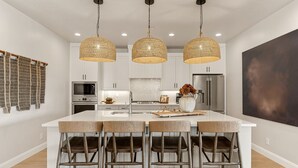 A modern kitchen with white cabinetry, a central island with bar stools, and three woven pendant lights. - Isle Be Back: Private Hot Tub + Rec Center Passes! (Washington)