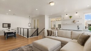 Una sala de estar moderna con un sofá color beige, una mesa de centro y una mesa de billar. Se ve una escalera con barandilla que conduce a un nivel superior.