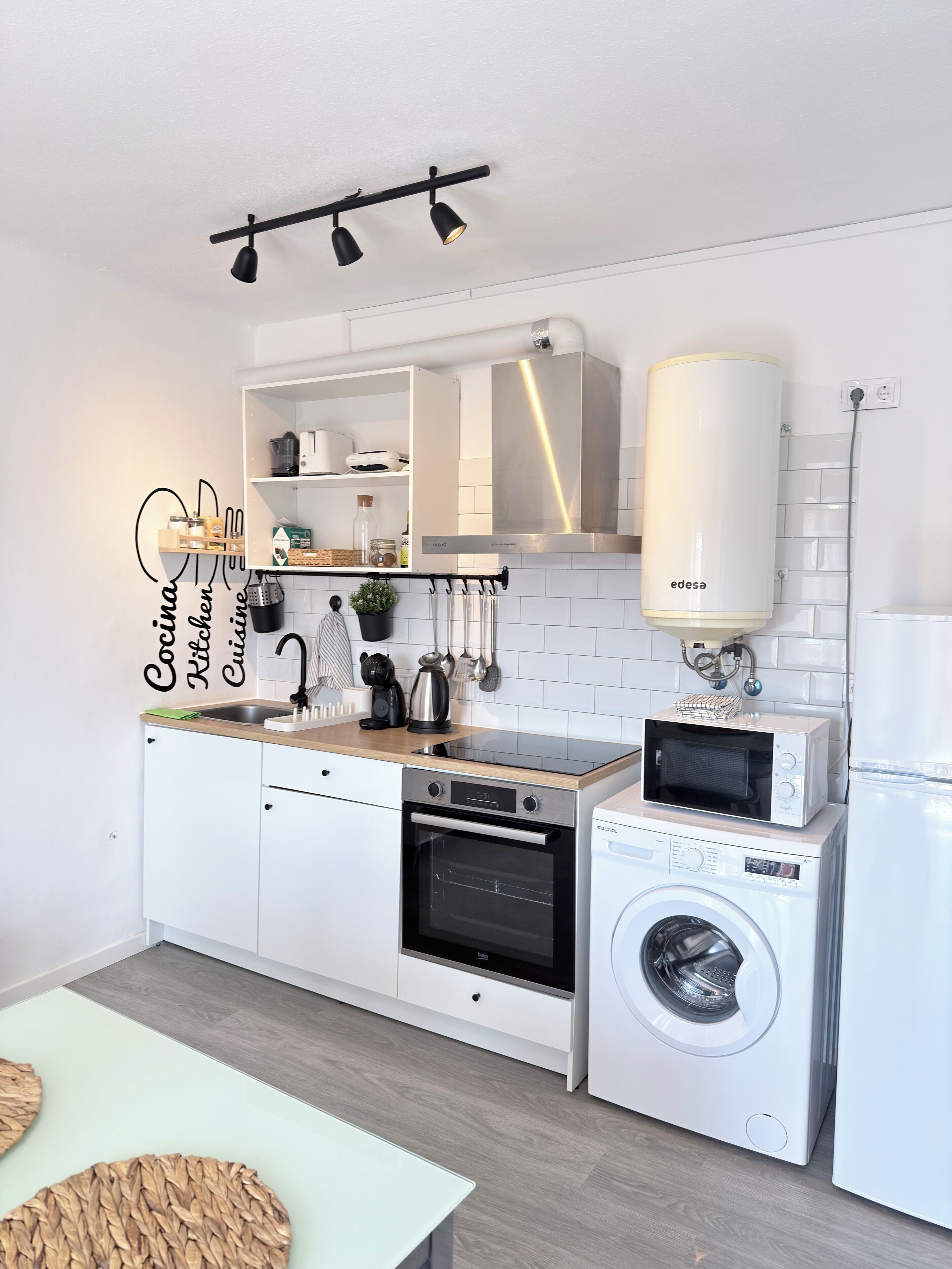 Una cocina moderna con armarios blancos, horno empotrado, microondas, lavadora y calentador de agua instalado en el techo.