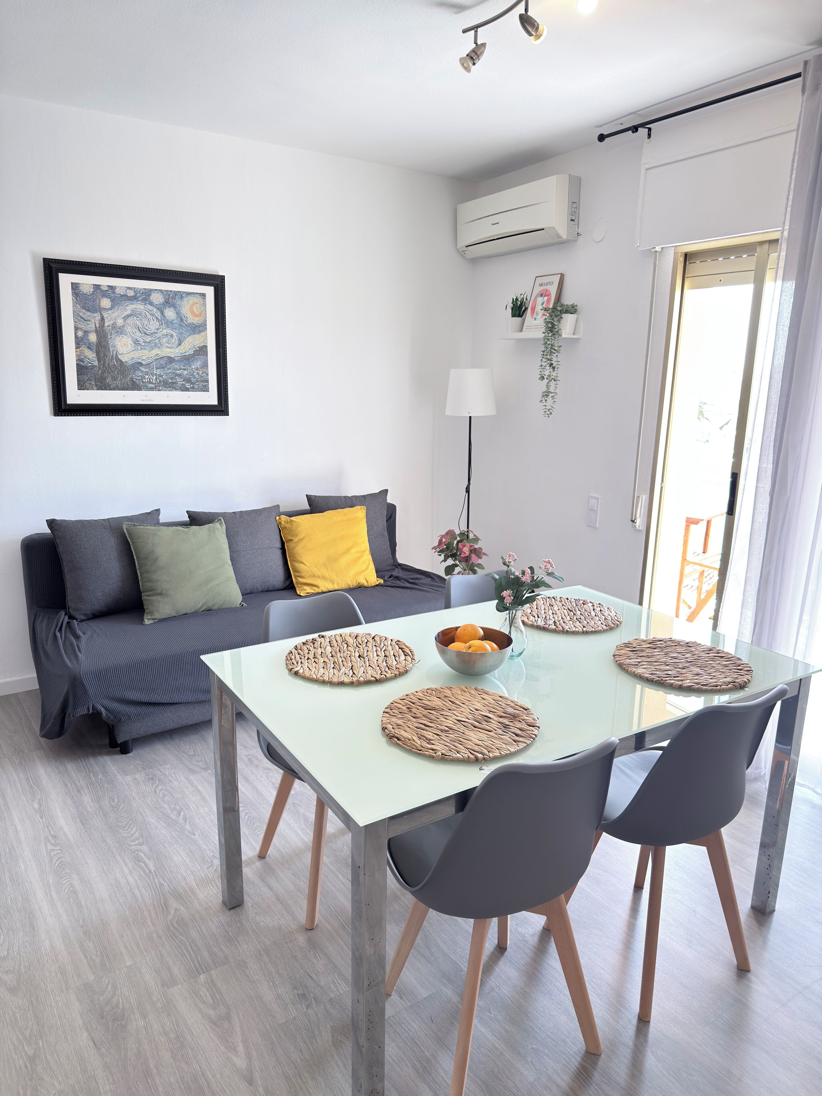 Una sala de estar moderna con una mesa de comedor de vidrio, un sofá gris y una obra de arte enmarcada en la pared.