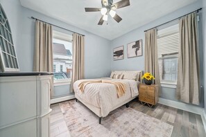 A bedroom with a bed, a nightstand with a vase, a ceiling fan, and a window with curtains.