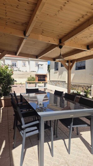 An outdoor patio with a table and chairs, a brick oven, and a covered area with wooden beams.