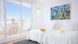 Une chambre d’hôtel avec un balcon, un lit avec du linge de lit blanc, un tableau au mur et un panier de fruits sur le lit.
