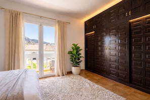 Une chambre à coucher avec un lit, une grande fenêtre avec des rideaux, une plante en pot et des portes en bois foncé.
