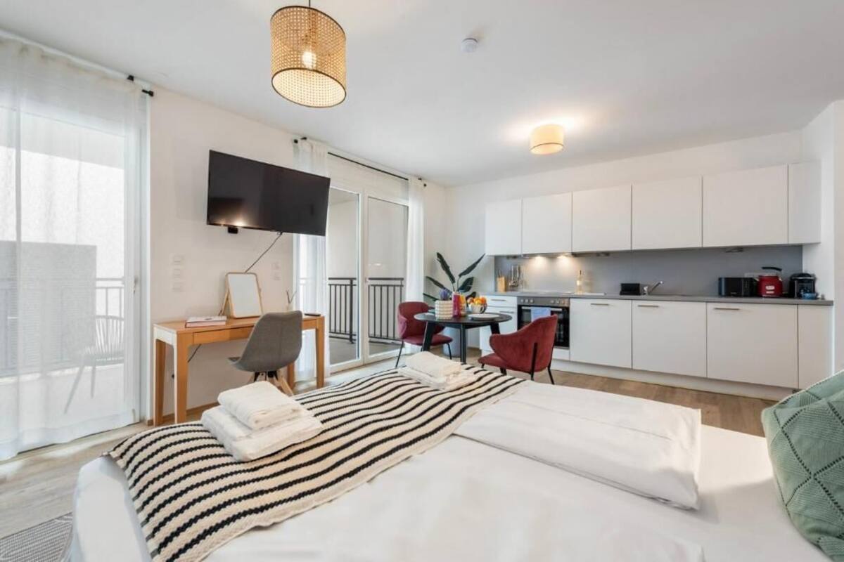 A modern hotel room with a bed, a desk, a TV, and a kitchenette.