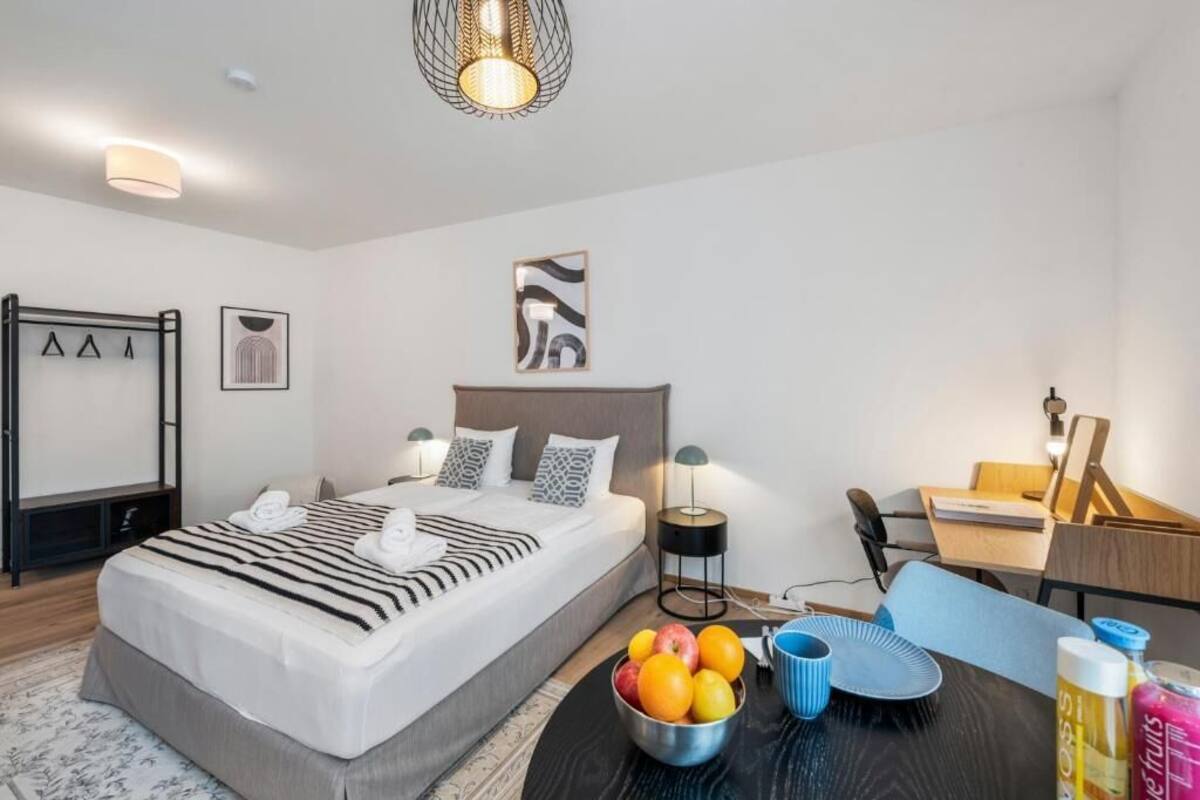 A modern hotel room with a neatly made bed, a small desk, a chair, and a fruit bowl on the side table.