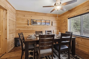 A wooden interior with a dining table set for four, a ceiling fan, and a window with a view of trees. - New Listing, Cozy and Rustic, Firepit, Hot Tub, and Free Attractions! (Gatlinburg)