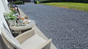 Un ensemble de chaise et de table en osier est installé sur une terrasse en gravier, agrémenté d’une plante en pot et d’une petite table sur laquelle se trouvent une tasse et une soucoupe.