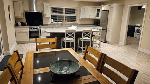 Une cuisine moderne avec des meubles blancs, un îlot central et un coin repas comprenant une table à plateau en verre et des chaises en bois.